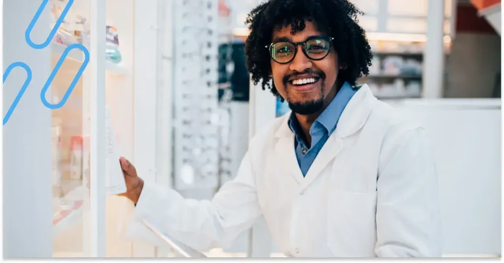 An image of a smiling pharmacist picking up a prescription from a shelf of prescriptions.