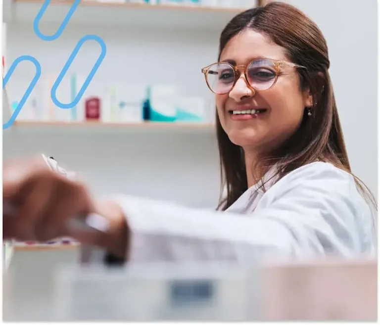 An image of a smiling pharmacist using a tool in the foreground of a shelf of prescriptions.