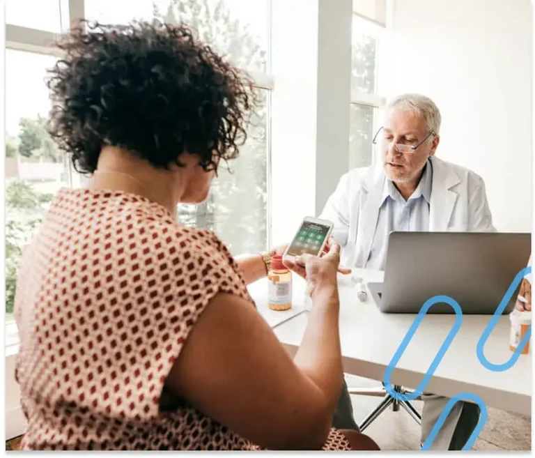 A lady on her phone conversing at a table with a doctor in front of his laptop.