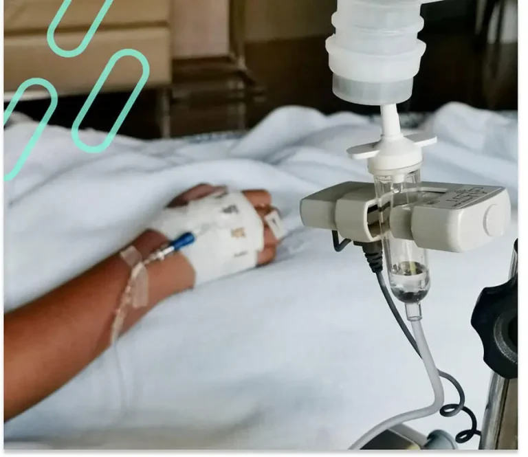 An image of an IV in the foreground with the patient's hand it is attached to in the background.