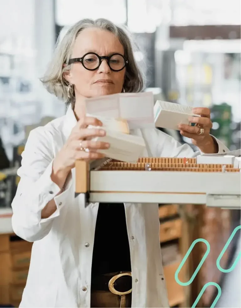 An image of a pharmacist inspecting a prescription note while holding a set of prescription boxes.