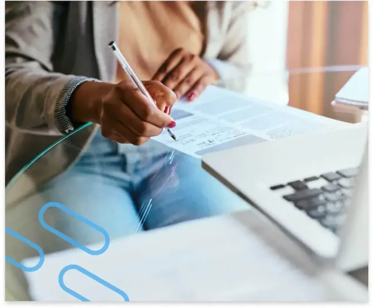 A person sitting at a round glass table manually filling out a form in front of their laptop.
